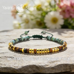May include: A beaded bracelet with turquoise, brown, and gold-colored beads. The bracelet is strung on a green cord and features the text "MORSE CODE: STRONG AS FUCK". The bracelet is displayed on a stone surface.