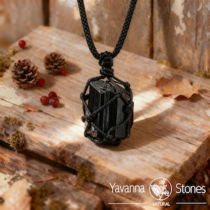 May include: A black tourmaline pendant necklace. The rectangular stone is encased in a woven black cord, suspended from a braided black cord. The necklace rests on a wooden surface with pinecones, berries, and the "Yavanna Stones" logo.