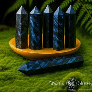 May include: Six black and blue crystal obelisks. Five obelisks stand upright in a wooden bowl, while one lies horizontally on a bed of green moss. The crystals have a dark blue, swirling pattern. The image includes the text "Yavanna Stones NATURAL."