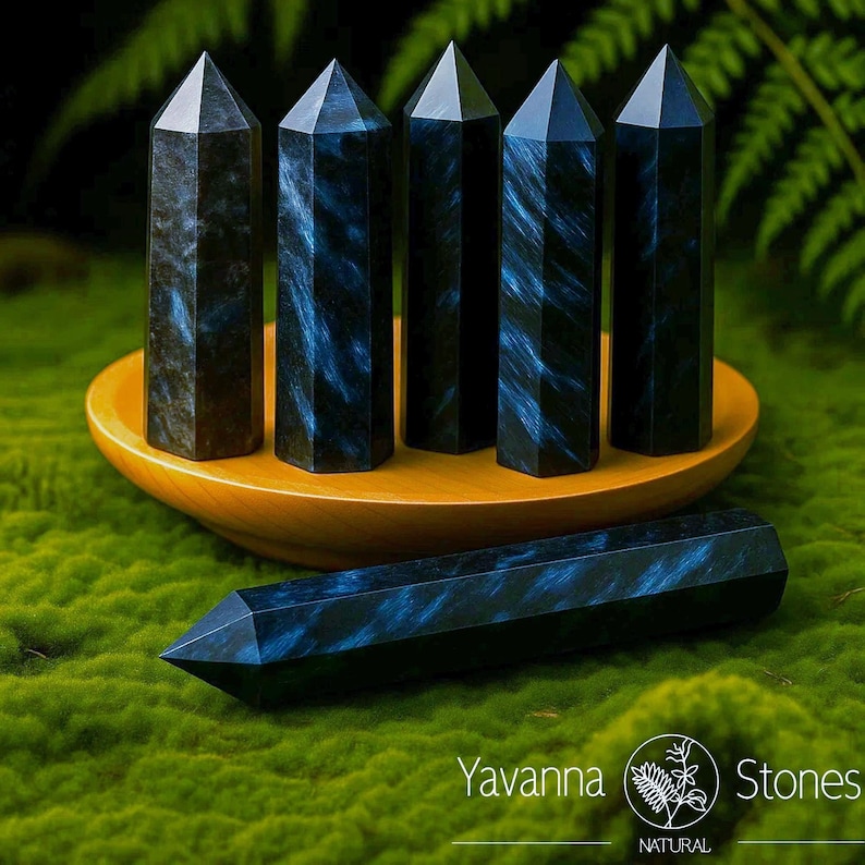 May include: Six black and blue crystal obelisks. Five obelisks stand upright in a wooden bowl, while one lies horizontally on a bed of green moss. The crystals have a dark blue, swirling pattern. The image includes the text "Yavanna Stones NATURAL."