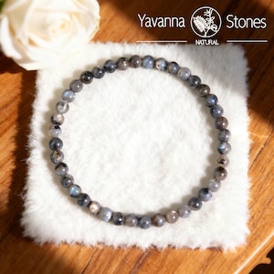 May include: A beaded bracelet made of small, round, gray and blue stones. The bracelet is displayed on a white, fluffy surface. The brand name "Yavanna Stones" is visible in the top right corner.