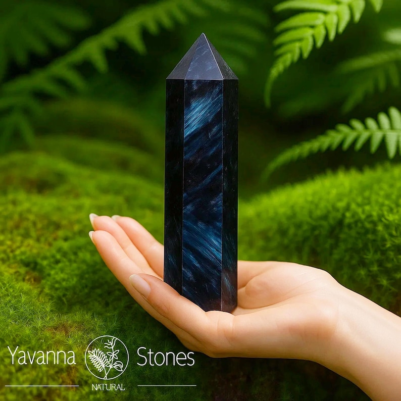 May include: A dark blue crystal obelisk with a pointed top, held in a hand against a backdrop of green moss and ferns. The crystal has a vertical striped pattern. The logo for "Yavanna Stones" is visible in the lower left corner.