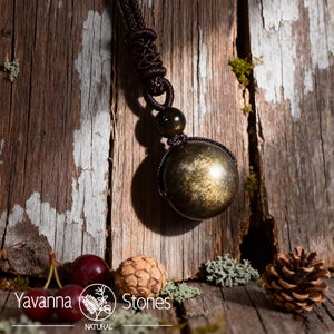 Pu&ograve; includere: Una collana con pendente a sfera in ossidiana dorata con un cordino marrone scuro. La sfera &egrave; racchiusa in un design marrone intrecciato. La collana &egrave; appesa su uno sfondo di legno rustico. Il logo "Yavanna Stones Natural" &egrave; visibile.