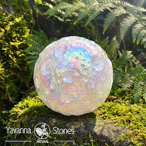 Op de afbeelding: Een heldere, iriserende bol met een textuur, die op een maan lijkt. De bol staat tegen een achtergrond van groen mos en varens. De bol heeft een regenboogglans. De tekst "Yavanna Stones NATURAL" staat onderaan.