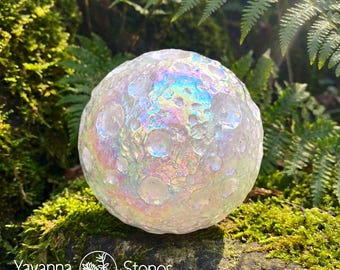Könnte beinhalten: Eine klare, irisierende Kugel mit strukturierter Oberfläche, die einem Mond ähnelt. Die Kugel befindet sich vor einem Hintergrund aus grünem Moos und Farnen. Die Kugel hat einen Regenbogen-Schimmer. Der Text "Yavanna Stones NATURAL" befindet sich unten.