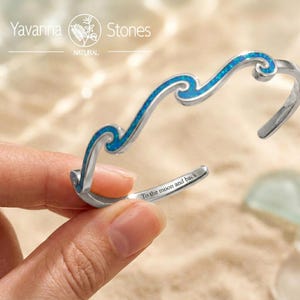 May include: A silver-toned cuff bracelet featuring a wave-shaped design inlaid with blue iridescent material. The inner surface of the metal band is engraved with the text To the moon and back. The brand name Yavanna Stones is visible.