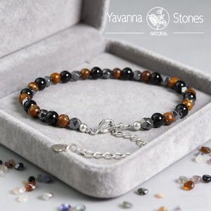 May include: A beaded bracelet with a mix of black, brown, and gray stones, displayed in a gray velvet box. The bracelet features a silver clasp and extender chain. The brand name "Yavanna Stones" is visible.