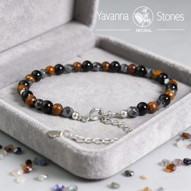 May include: A beaded bracelet with a mix of black, brown, and gray stones, displayed in a gray velvet box. The bracelet features a silver clasp and extender chain. The brand name "Yavanna Stones" is visible.