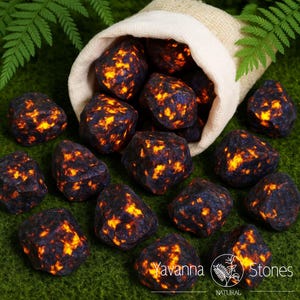 Peut inclure: Plusieurs pierres bleu foncé et orange sortent d'un sac en lin beige. Les pierres ont un aspect texturé avec des motifs orange et jaune vifs. L'image est posée sur un fond vert, avec le texte "Yavanna Stones NATURAL" visible.