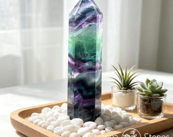May include: A tall, polished fluorite crystal tower featuring distinct bands of deep purple, emerald green, and translucent white. The hexagonal point stands upright among smooth white river stones on a light wooden tray with two small potted succulents.