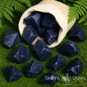 Puede incluir: Piedras facetadas de color azul oscuro con inclusiones brillantes, algunas de ellas saliendo de una bolsa de cord&oacute;n beige. Las piedras est&aacute;n dispuestas sobre una superficie verde, con hojas de helecho en el fondo. La marca "Yavanna Stones" es visible.