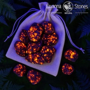 May include: A collection of polished stones with a fiery orange and dark brown pattern, spilling from a beige drawstring bag. The bag is set against a dark green background with fern leaves. The text "Yavanna Stones NATURAL" is visible in the upper right corner.
