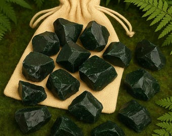 Puede incluir: Una colección de piedras preciosas facetadas de color verde oscuro esparcidas alrededor de una bolsa beige con cordón. Las piedras preciosas tienen una textura natural rugosa y formas variadas. El fondo es una superficie verde oscuro con hojas de helecho.