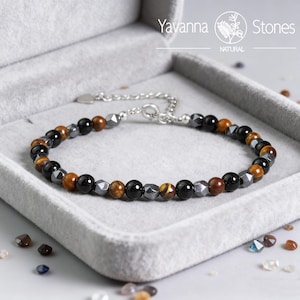 May include: A beaded bracelet with a mix of black, brown, and gray stones. The bracelet is on a gray velvet surface inside a gray box. The bracelet has a silver chain and clasp. The brand name "Yavanna Stones" is visible.