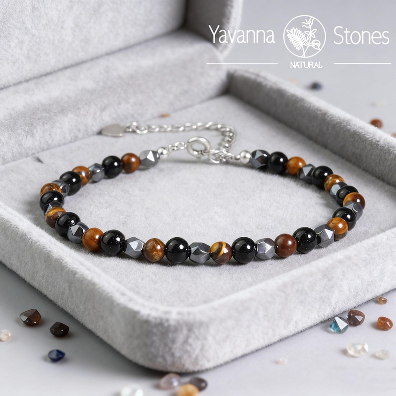 May include: A beaded bracelet with a mix of black, brown, and gray stones. The bracelet is on a gray velvet surface inside a gray box. The bracelet has a silver chain and clasp. The brand name "Yavanna Stones" is visible.