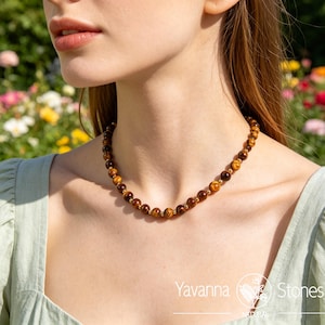 May include: A beaded necklace featuring tiger's eye stones and gold-colored accents. The necklace is worn around the neck. The background is a blurred view of a garden with colorful flowers. The necklace is from Yavanna Stones.