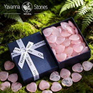 May include: A navy blue gift box tied with a white ribbon printed with "Crystal Jewelry". The box is open, revealing a collection of pink heart-shaped stones. The stones are scattered around the box on a bed of green moss. The top left corner of the image has the text "Yavanna Stones".