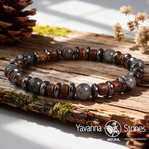 May include: A beaded bracelet featuring a mix of natural stone beads in shades of gray, brown, and black. The bracelet is strung on a durable cord and is displayed on a rustic wooden surface. The bracelet includes the text "Yavanna Stones".