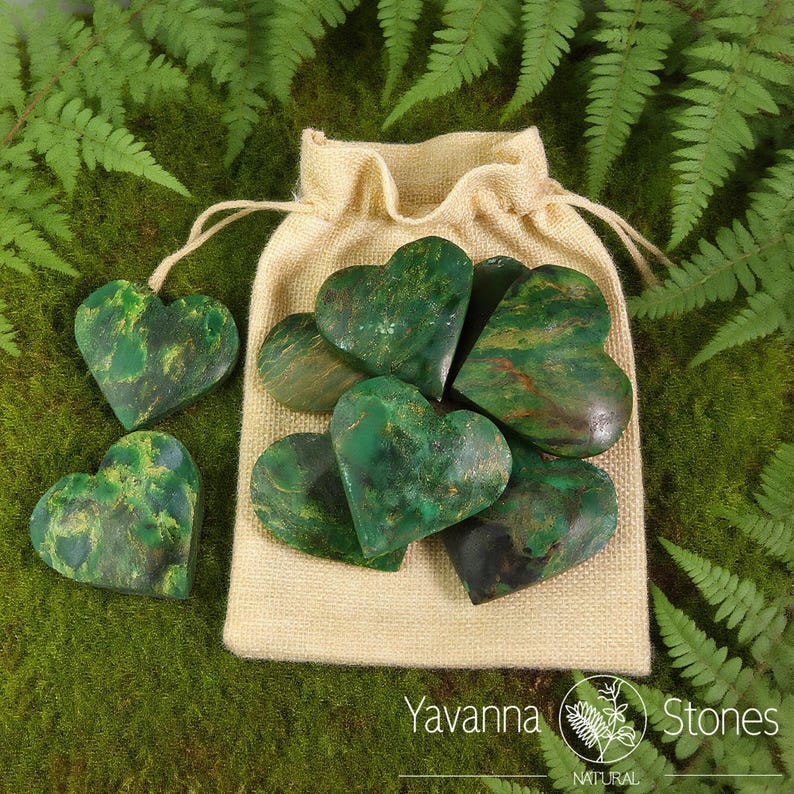 May include: A collection of green heart-shaped stones with natural patterns, some outside a beige drawstring bag. The stones have a mix of green and brown hues. The background features green moss and fern leaves. The text "Yavanna Stones NATURAL" is visible.