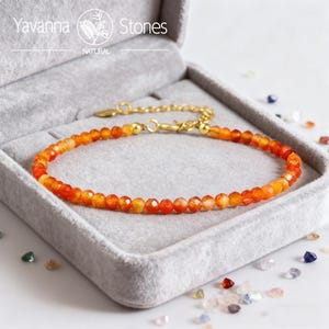 May include: An orange gemstone bracelet with a gold-colored clasp and chain, presented in a gray velvet box. The bracelet is made of small, faceted orange beads. The brand name "Yavanna Stones" is visible in the top left corner.