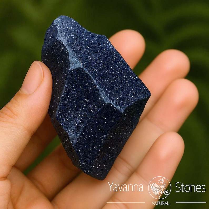 Puede incluir: Una gema azul oscuro, tallada en bruto, con inclusiones brillantes, se sostiene en una mano. La piedra tiene una superficie facetada y una forma natural. El fondo es verde borroso. La piedra est&aacute; etiquetada como "Yavanna Stones".