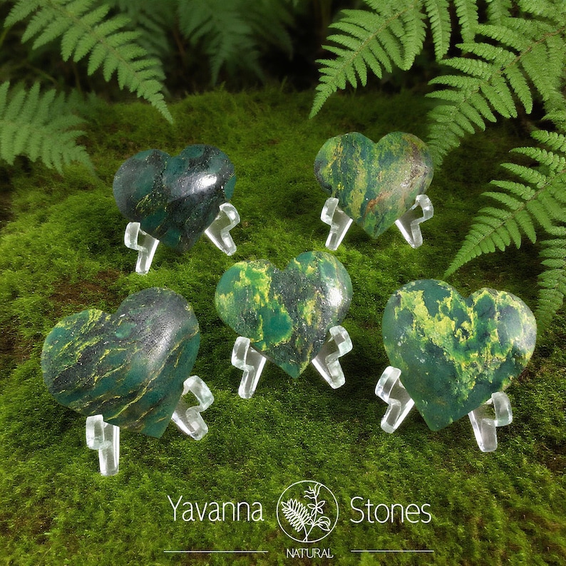 May include: Five heart-shaped stones in shades of green and yellow, each displayed on a clear stand. The stones are set against a backdrop of green moss and fern leaves. The logo "Yavanna Stones NATURAL" is visible.