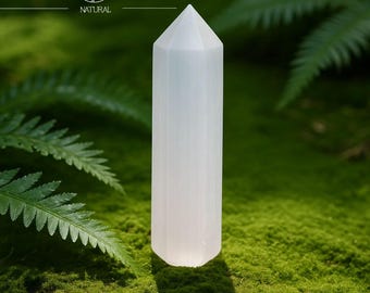 May include: A white selenite crystal tower stands on a bed of green moss, with fern leaves in the background. The crystal is faceted and has a pointed top. The image includes the text "Yavanna Stones NATURAL".