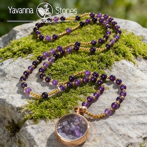 Può includere: Una collana con perline di ametista e dettagli color oro. La collana include un grande pendente rotondo con una cornice color oro rosa, riempito di pietre di ametista. La collana è esposta su muschio verde.