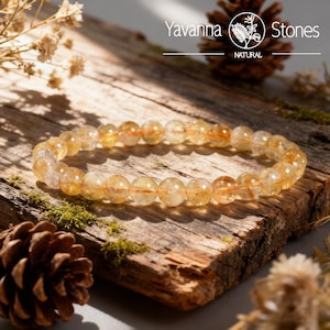 May include: A beaded bracelet made of golden-yellow citrine stones. The bracelet is displayed on a rustic wooden surface with a pine cone and sprigs of dried flowers. The brand name "Yavanna Stones" is visible in the background.