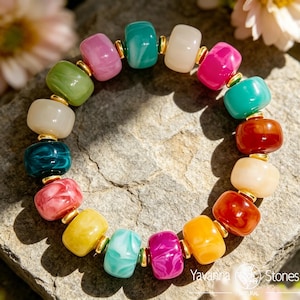 May include: A colorful beaded bracelet featuring a variety of vibrant, rounded beads in shades of pink, green, teal, yellow, and orange. The beads are separated by small gold-colored spacers. The bracelet is displayed on a textured stone surface.