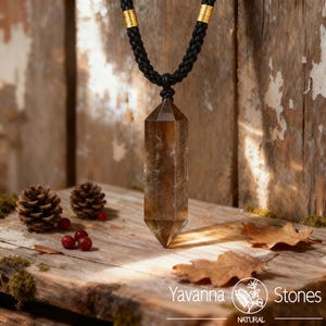 May include: A brown crystal pendant necklace with a black braided cord and gold accents. The crystal is a long, hexagonal shape. The necklace is displayed on a wooden surface with pinecones, berries, and leaves. The brand name "Yavanna Stones" is visible.