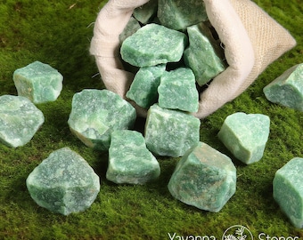 May include: A collection of rough, green aventurine stones spilling from a beige burlap bag onto a bed of green moss. The stones vary in size and shape, with a natural, textured appearance. The image is well-lit, highlighting the stones' color and details.