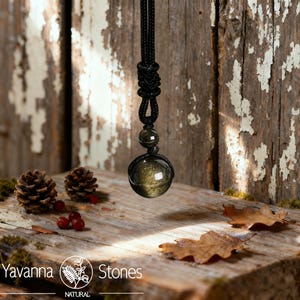 May include: A black cord necklace with a large, round, golden-brown gemstone pendant. The pendant is suspended from a knotted section of the cord. The image also includes pine cones, berries, and fallen leaves on a wooden surface. The brand name "Yavanna Stones" is visible.