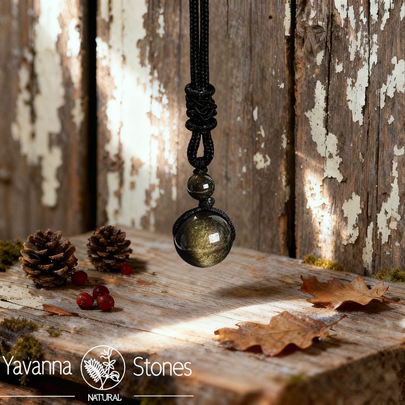 Pu&ograve; includere: Una collana con cordino nero con un grande pendente sferico in pietra preziosa marrone dorato. Il pendente &egrave; sospeso a una sezione annodata del cordino. L'immagine include anche pigne, bacche e foglie cadute su una superficie di legno. Il marchio "Yavanna Stones" &egrave; visibile.