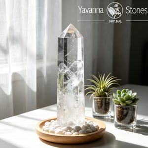 May include: A clear, faceted quartz crystal point standing upright in a round wooden tray filled with small white stones. The text Yavanna Stones Natural is visible in the upper right corner. Two small glass pots containing green plants sit nearby.