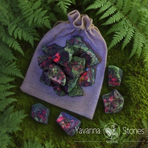 May include: A collection of rough-cut stones with green and red hues, spilling from a drawstring bag. The stones are scattered on a bed of green moss and ferns. The bag is a light purple color. The logo "Yavanna Stones" is visible.