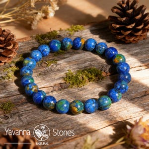 May include: A beaded bracelet featuring round, blue and green stones. The bracelet is displayed on a rustic wooden surface with moss and pinecones. The brand name "Yavanna Stones" is visible in the lower left corner.