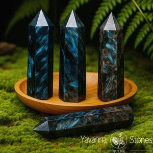 May include: Four blue-black crystal obelisks with a natural, patterned appearance. Three stand upright in a wooden bowl, while one lies horizontally. The crystals have a polished, faceted top. The image includes the text "Yavanna Stones".