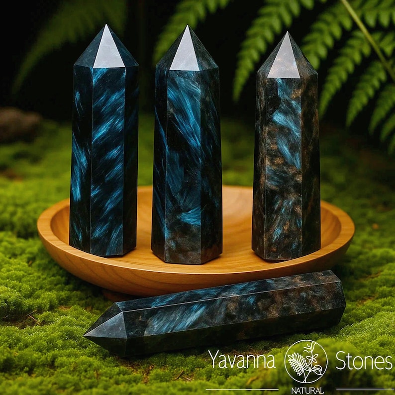 May include: Four blue-black crystal obelisks with a natural, patterned appearance. Three stand upright in a wooden bowl, while one lies horizontally. The crystals have a polished, faceted top. The image includes the text "Yavanna Stones".