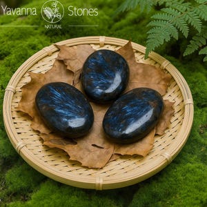 May include: Three polished, oval stones with a dark blue and black pattern, presented on a woven bamboo tray with dried leaves. The tray rests on green moss. The text "Yavanna Stones NATURAL" is visible.