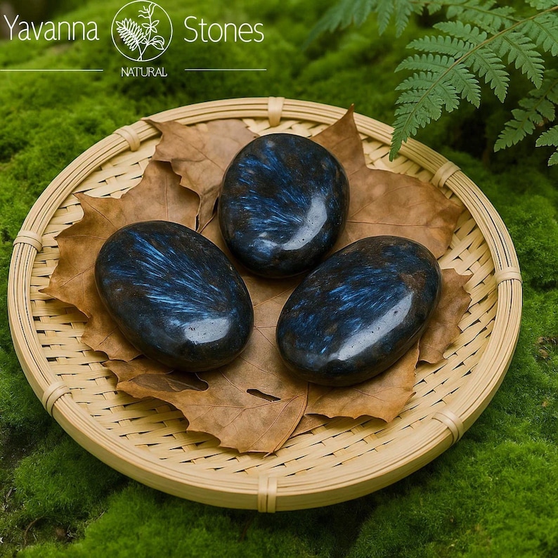 May include: Three polished, oval stones with a dark blue and black pattern, presented on a woven bamboo tray with dried leaves. The tray rests on green moss. The text "Yavanna Stones NATURAL" is visible.