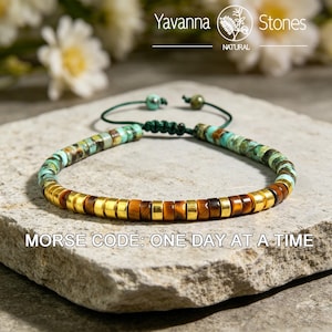 May include: A beaded bracelet with turquoise, brown, and gold-colored beads. The bracelet is strung on a green cord and features the text "MORSE CODE: ONE DAY AT A TIME". The bracelet is displayed on a stone surface.