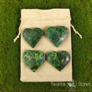 May include: Four heart-shaped green stones with yellow veining are arranged on a beige drawstring bag. The stones are polished and have a natural, organic appearance. The bag is on a green surface. The text "Yavanna Stones NATURAL" is visible.