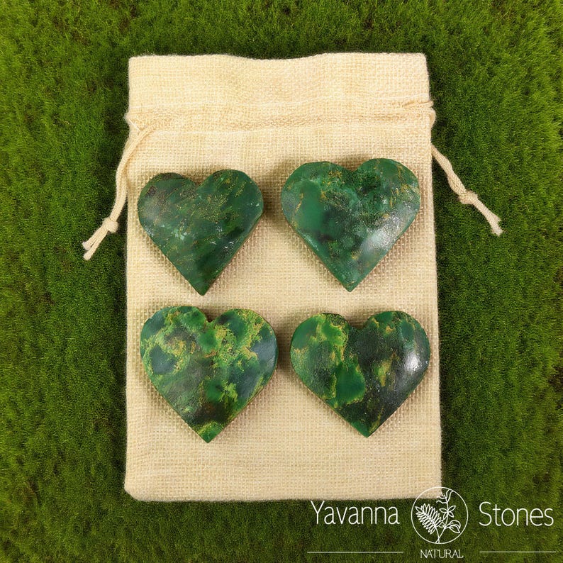 May include: Four heart-shaped green stones with yellow veining are arranged on a beige drawstring bag. The stones are polished and have a natural, organic appearance. The bag is on a green surface. The text "Yavanna Stones NATURAL" is visible.