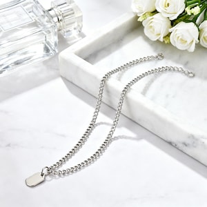 May include: A silver-colored necklace with a chain design and a small, rectangular pendant. The necklace is displayed on a white marble tray, with a perfume bottle and white roses in the background. The chain is approximately 18 inches long.