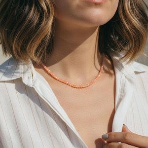 May include: A delicate necklace composed of small, peach-colored beads. The necklace is worn against a white collared shirt. The beads appear to be evenly sized and strung together to form a simple, elegant piece of jewelry.