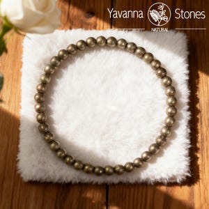 May include: A beaded bracelet made of small, round, metallic pyrite stones. The bracelet is displayed on a fluffy, white surface. The brand name "Yavanna Stones" is visible in the top left corner of the image.