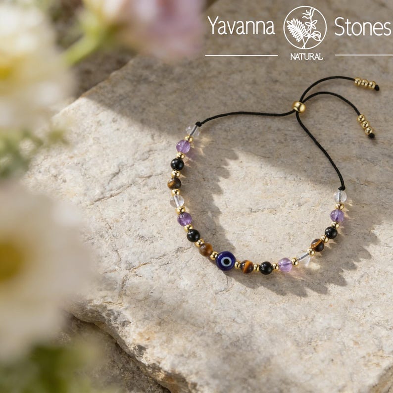 May include: A beaded bracelet featuring a variety of natural stones, including tiger's eye, amethyst, and black onyx. The bracelet includes a blue evil eye bead and gold accents. The bracelet is on a black cord with an adjustable closure.