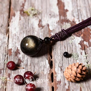 Pu&ograve; includere: Una collana con pendente in pietra preziosa nera e oro con un cordoncino intrecciato. Il pendente &egrave; una pietra rotonda e lucida con riflessi dorati. La collana &egrave; esposta su una superficie di legno rustico con piccole bacche rosse e una pigna.