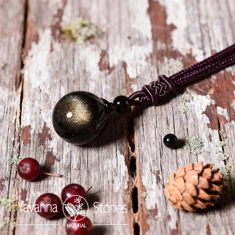 Pu&ograve; includere: Una collana con pendente in pietra preziosa nera e oro con un cordoncino intrecciato. Il pendente &egrave; una pietra rotonda e lucida con riflessi dorati. La collana &egrave; esposta su una superficie di legno rustico con piccole bacche rosse e una pigna.